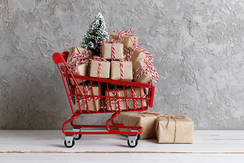 Miniature red shopping cart filled with kraft-wrapped gifts and a small snowy tree on a wooden floor against a textured wall