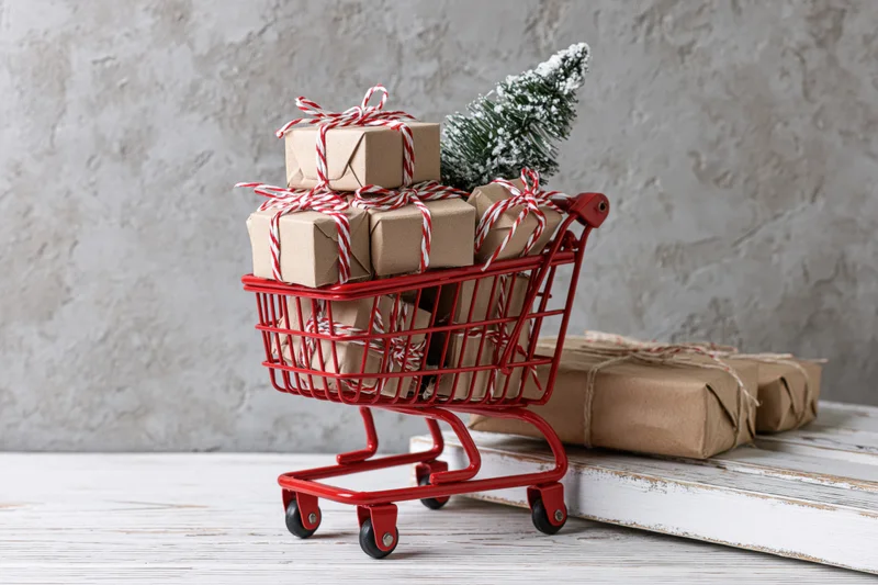 Miniature red shopping cart filled with kraft-wrapped gifts and a small snowy tree on a wooden surface