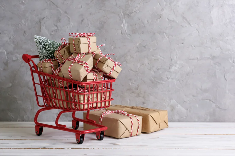 Miniature red shopping cart overflowing with kraft-wrapped gifts on wooden floor against textured wall