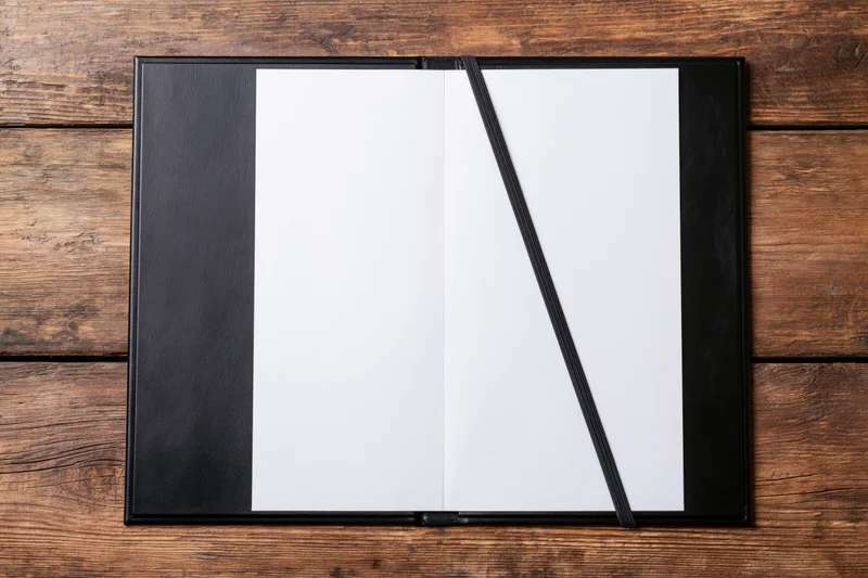 Open black leather restaurant menu presenter with blank white pages on wooden table