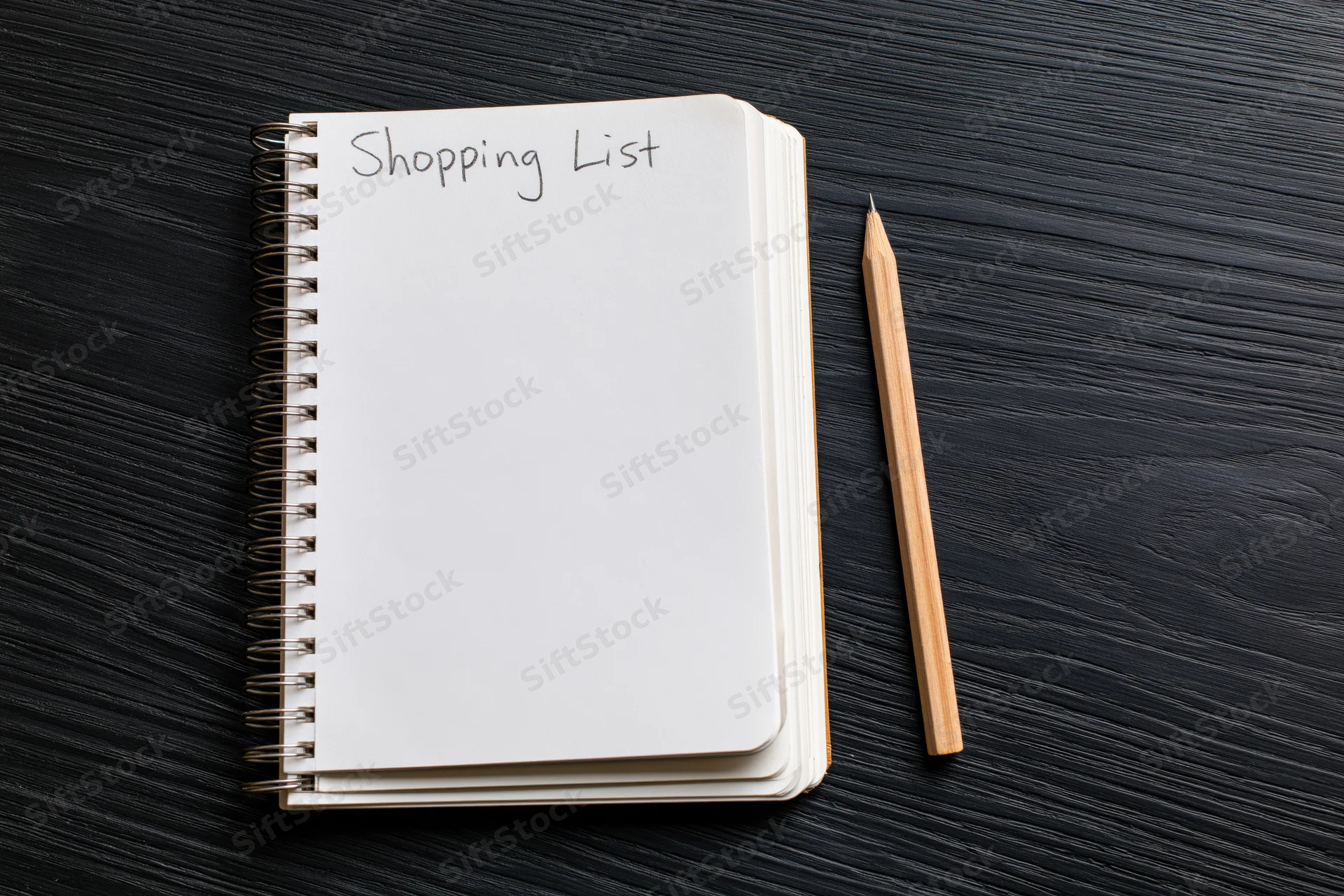 Spiral notepad with shopping list handwritten at top on dark wood surface next to pencil