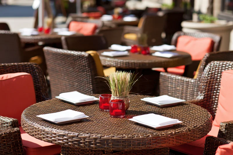 Round wicker table set for dining on an outdoor restaurant terrace with orange cushions and centerpiece