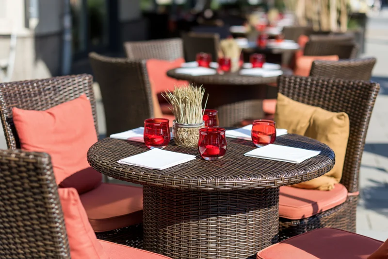 Round wicker tables set for dining on an outdoor restaurant terrace with orange cushions and red glasses