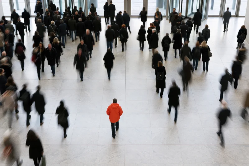 Overhead view of a crowd of people walking through a modern lobby with one person in an orange hoodie standing out in the center