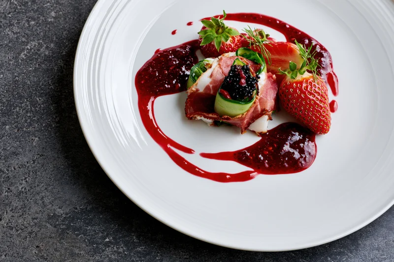 Fine dining appetizer with bacon-wrapped cucumber, strawberries, blackberries, and red sauce on a white plate over dark surface
