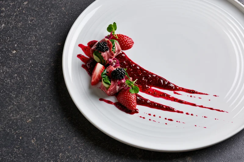 Fine dining appetizer with berry sauce and fruit garnish on white plate over dark surface