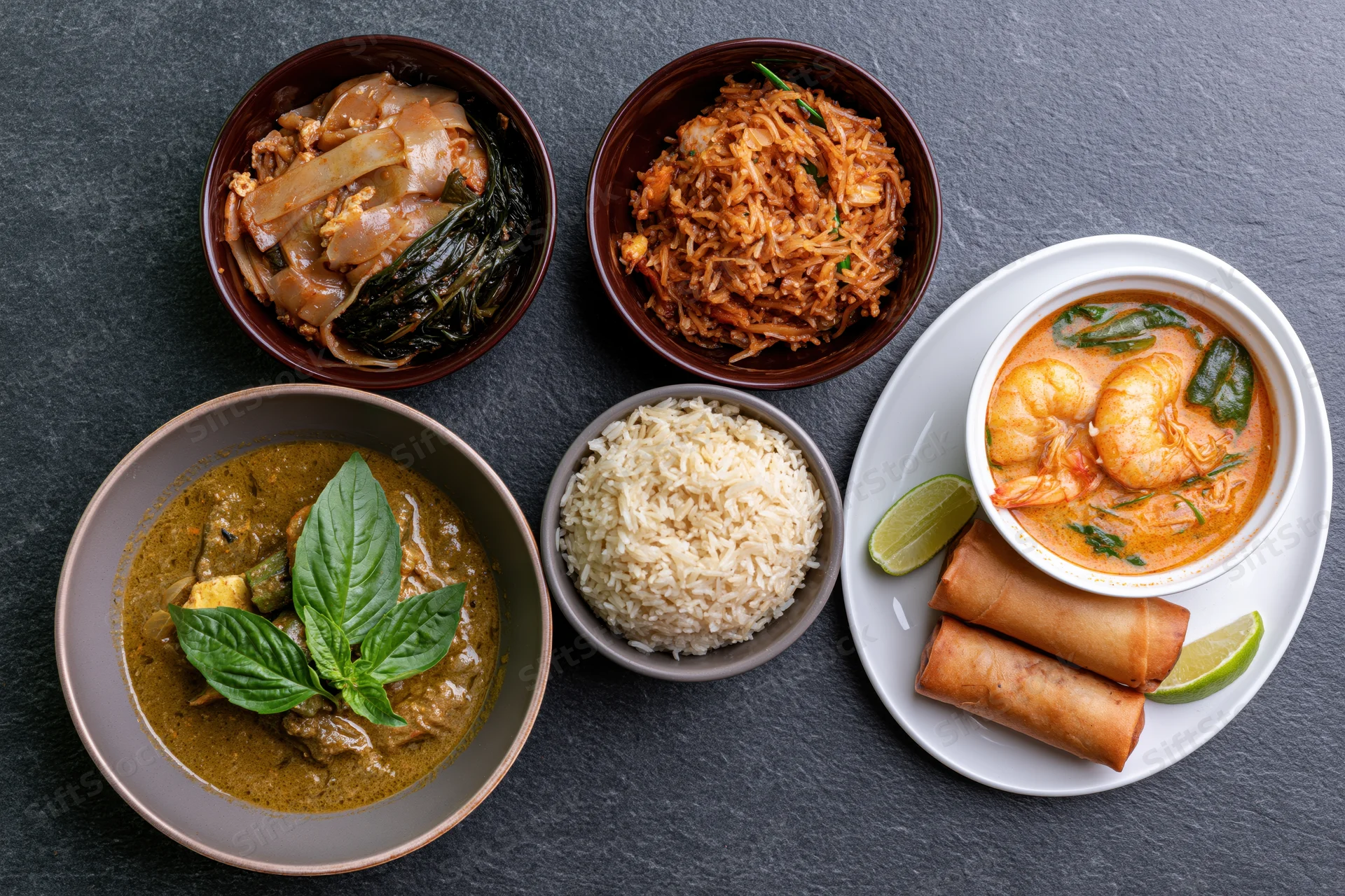 Overhead flat lay of thai dishes including curry, rice, noodles, shrimp soup, and spring rolls arranged on dark slate surface