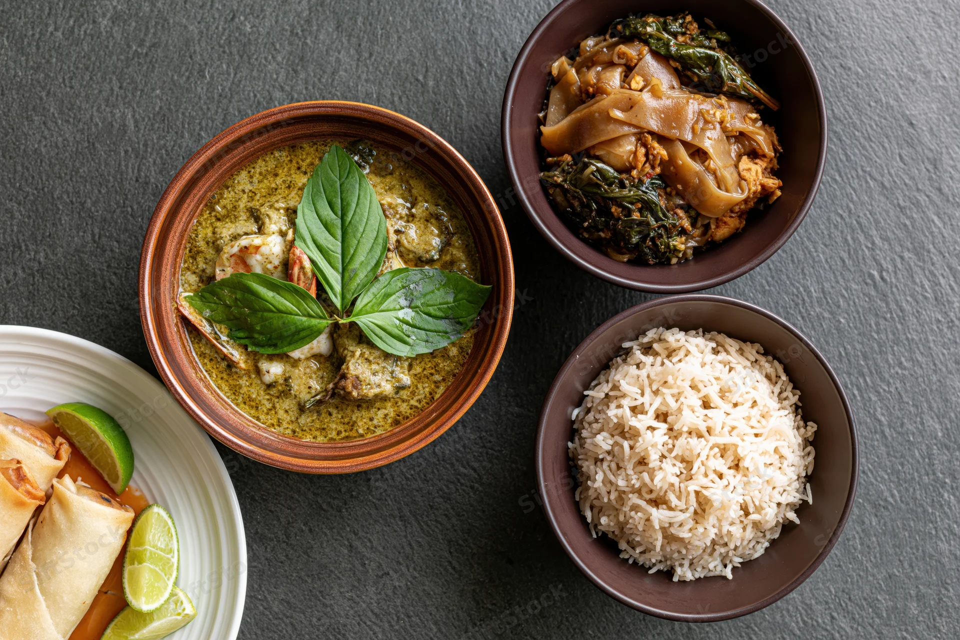 Overhead flat lay of four thai dishes arranged on dark slate surface