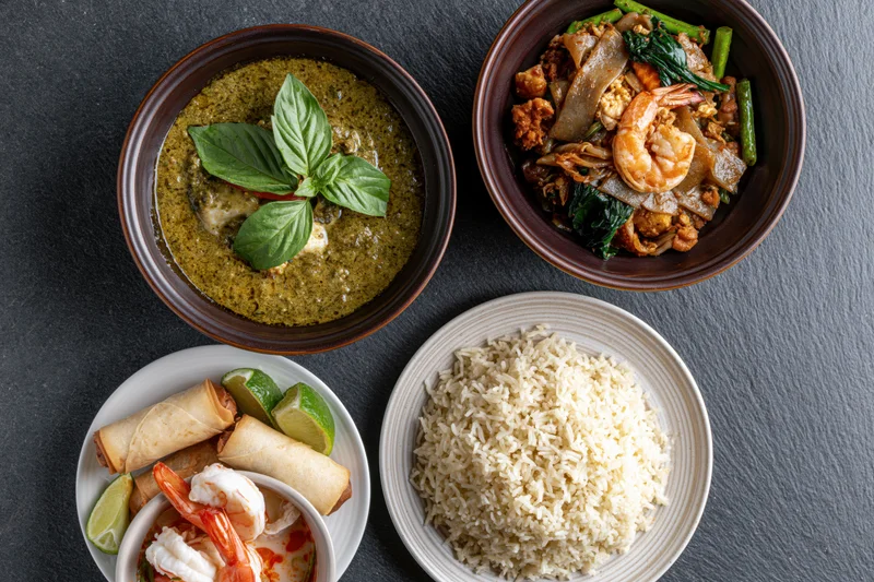 Four thai dishes arranged overhead on dark slate surface including green curry, pad thai, rice, and spring rolls with lime garnish