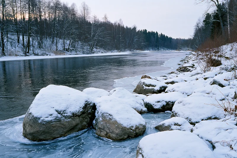 Partially frozen river winding through snow-covered forested landscape at twilight