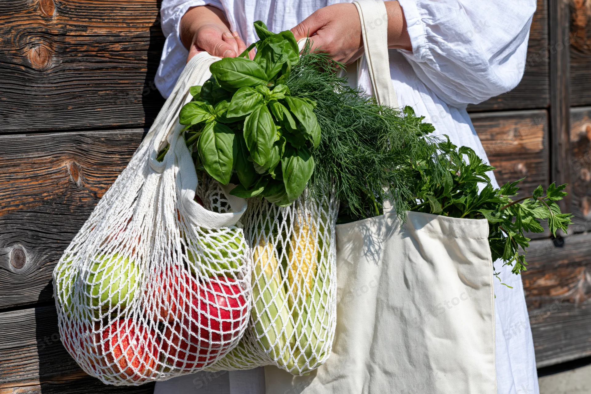 Woman holding reusable shopping bags filled with fresh produce and herbs outdoors