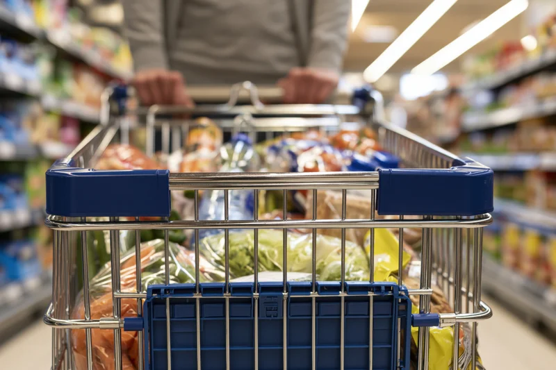 Person pushing a metal shopping cart filled with groceries in a supermarket aisle