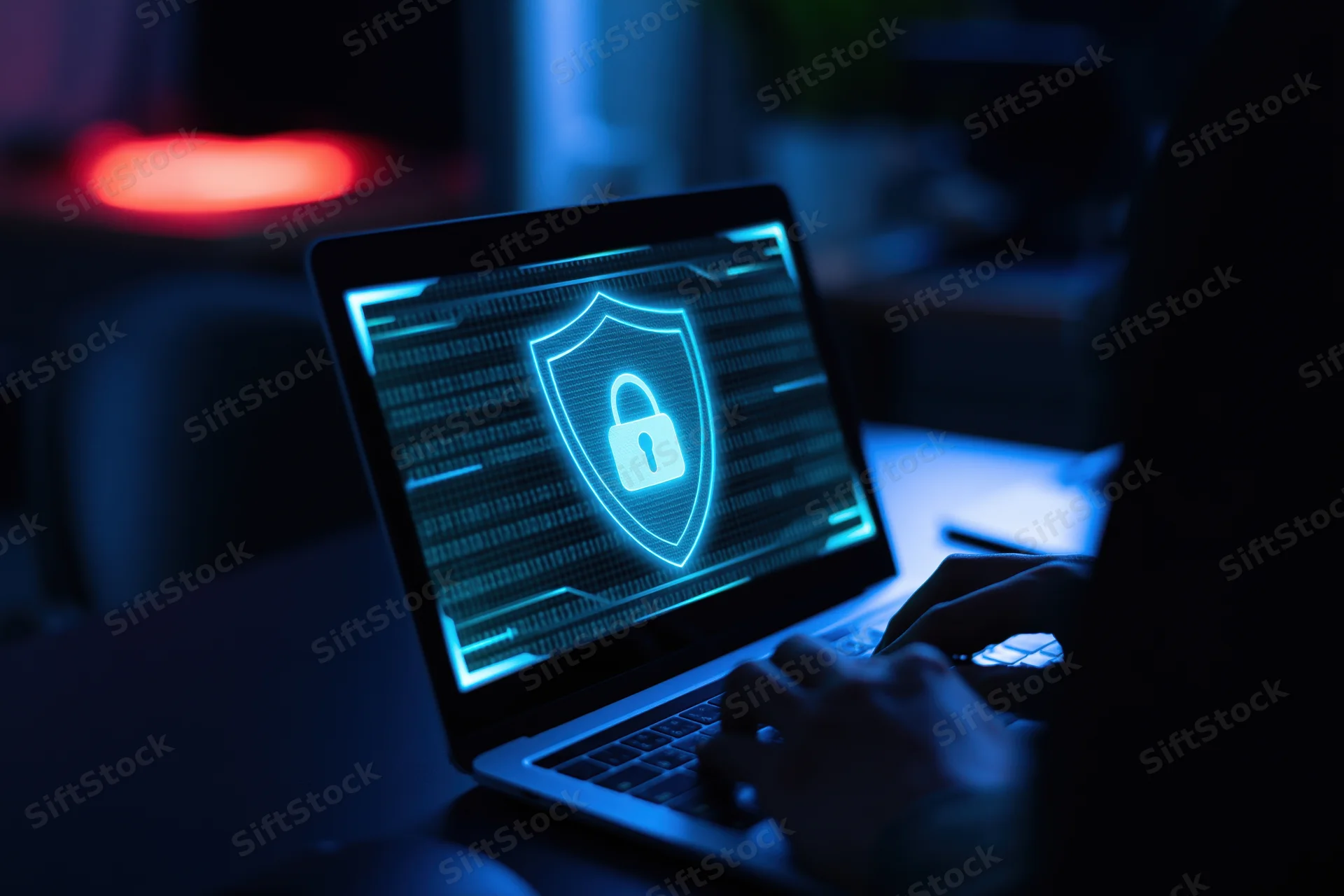 Person typing on laptop with glowing shield and lock on screen in dark office