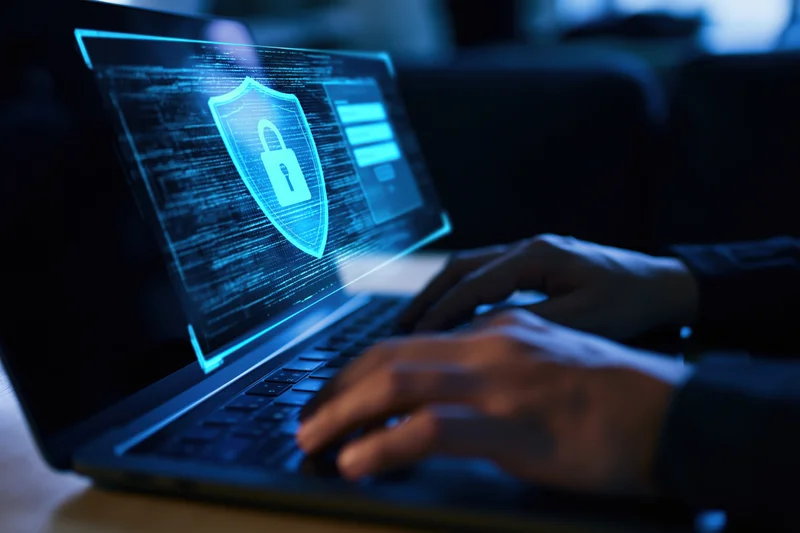 Hands typing on a laptop keyboard with a glowing shield and lock symbol on the screen in a dark room