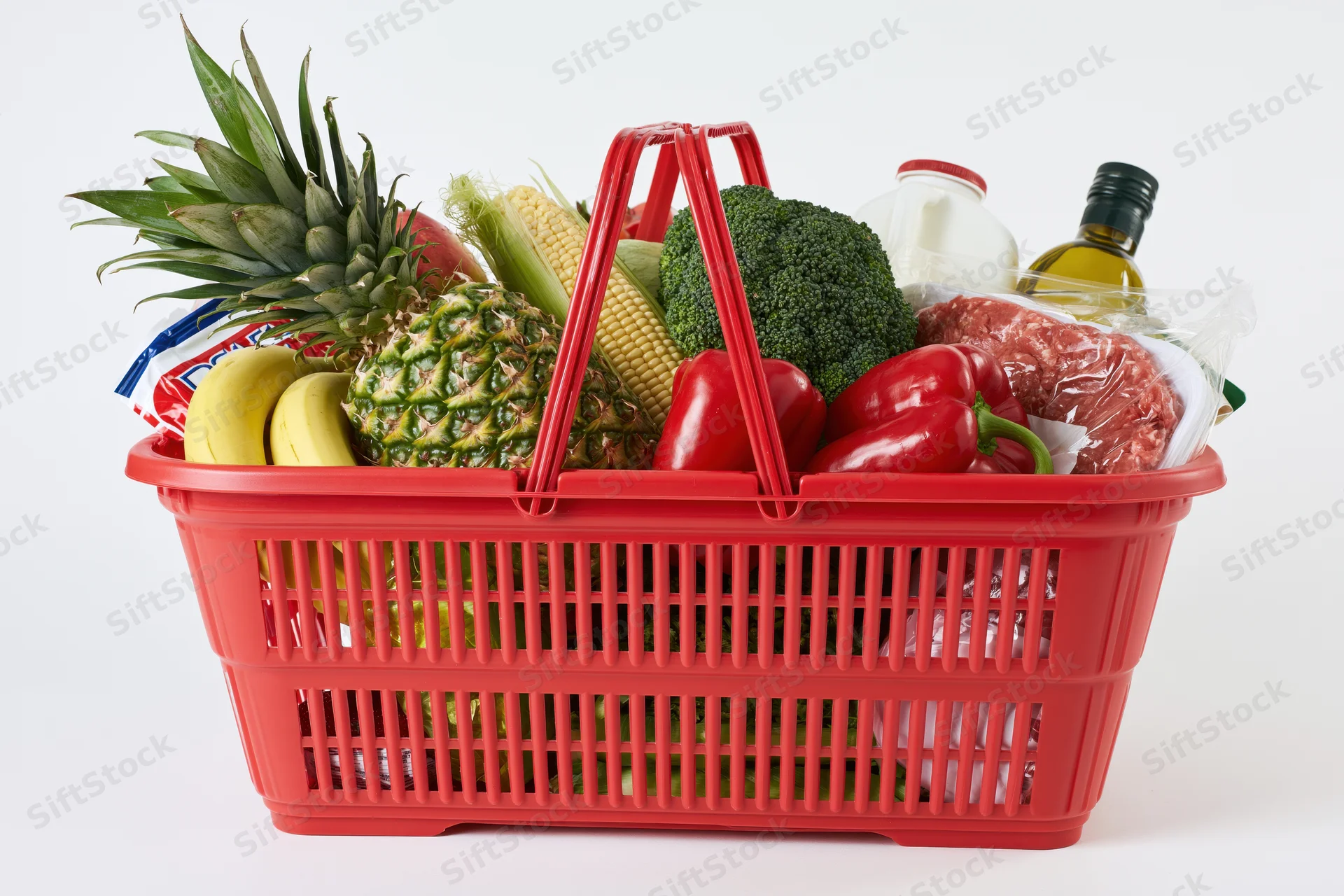 Red plastic shopping basket filled with fresh groceries including fruit vegetables meat and dairy against white background