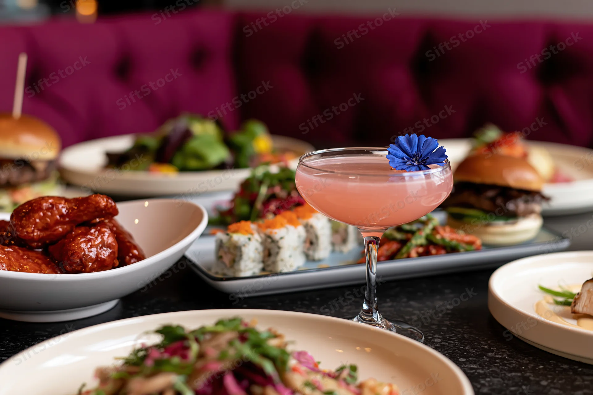 Pink cocktail garnished with blue flower on a restaurant table surrounded by various dishes including sushi, burgers, and glazed wings