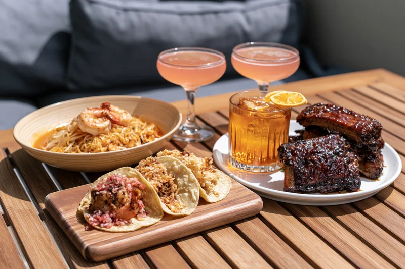 Restaurant table spread with shrimp noodles, crispy tacos, ribs, and cocktails on a wooden patio table