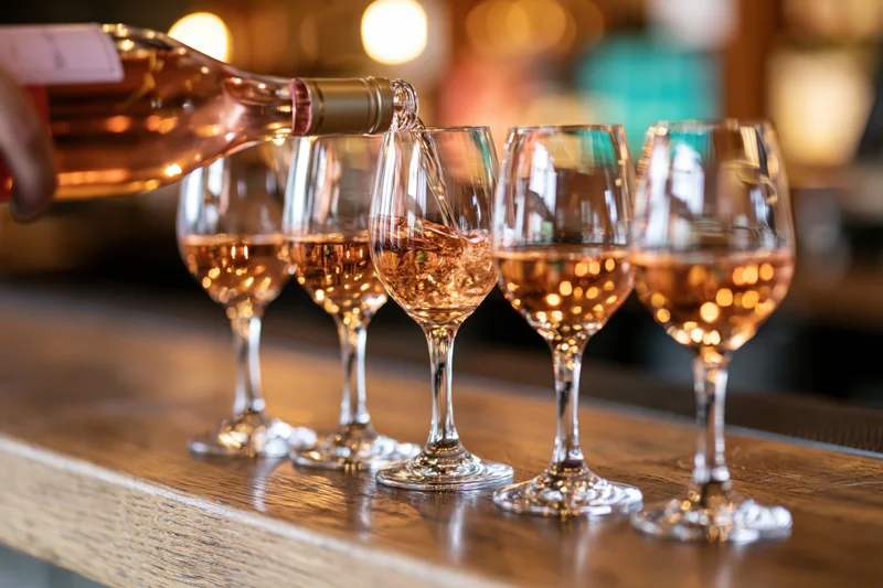 Rosé wine being poured into a row of five glasses on a wooden bar counter