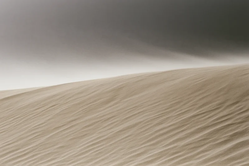 Wind blowing sand off the crest of a desert dune under an overcast sky