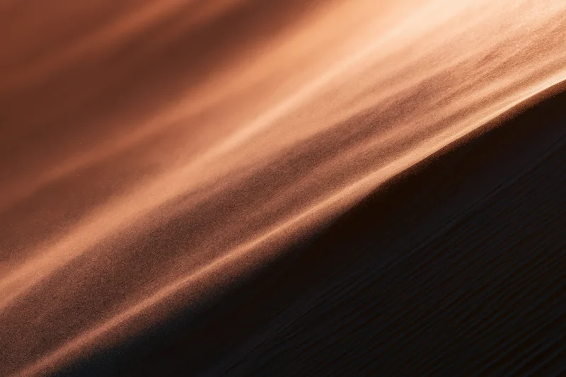 Fine sand grains blowing off a sharp ridge of a desert dune in warm light