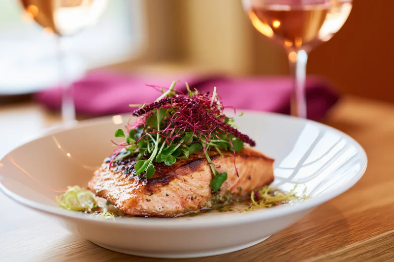 Seared salmon fillet topped with microgreens and red amaranth on a white plate at a restaurant table
