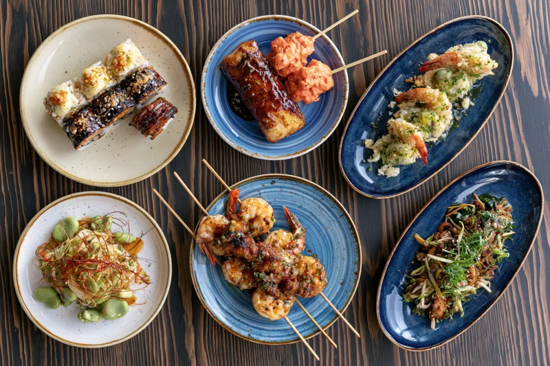 Seven restaurant dishes arranged on dark wooden table with sushi, shrimp skewers, tempura, salad and garnishes on ceramic plates