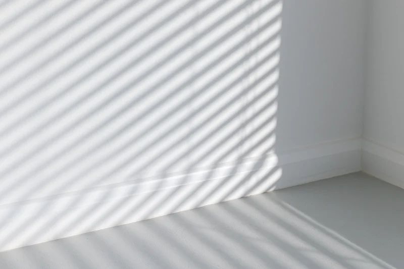 Diagonal shadow stripes from window blinds across white wall and floor in empty room corner