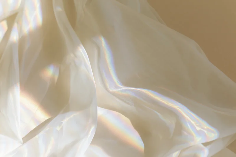 Sheer white fabric draped loosely across a pale beige wall with rainbow light patterns from sunlight diffraction