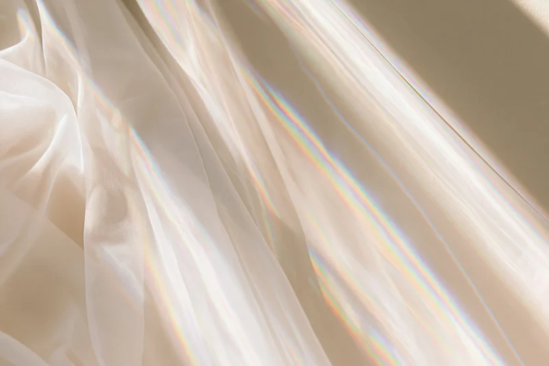 Sheer white fabric draped loosely across a pale beige wall with soft sunlight creating iridescent rainbow reflections