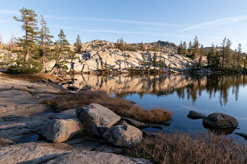 Alpine tarn reflecting granite peaks and scattered conifer trees under clear blue sky