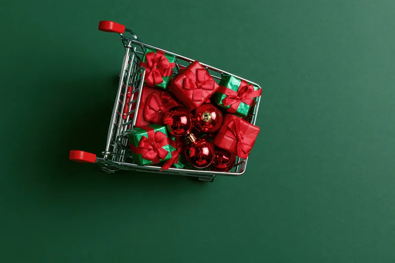 Miniature shopping cart filled with wrapped gifts and red ornaments on green surface