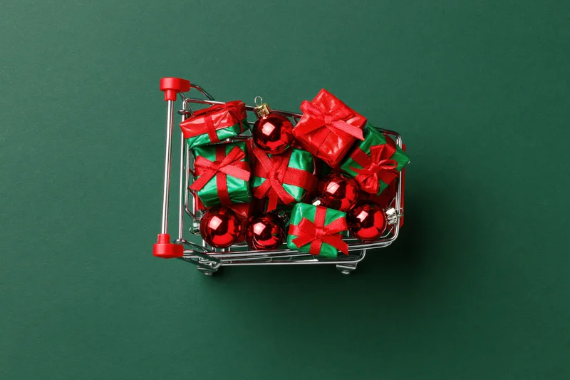Miniature shopping cart filled with red and green wrapped gifts and shiny red ornaments on a green background