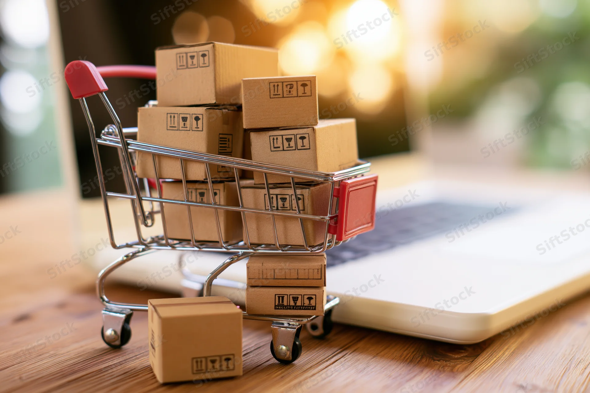 Miniature shopping cart filled with cardboard boxes on a desk beside a laptop