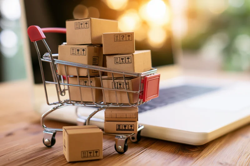 Miniature shopping cart filled with cardboard boxes on a desk beside a laptop