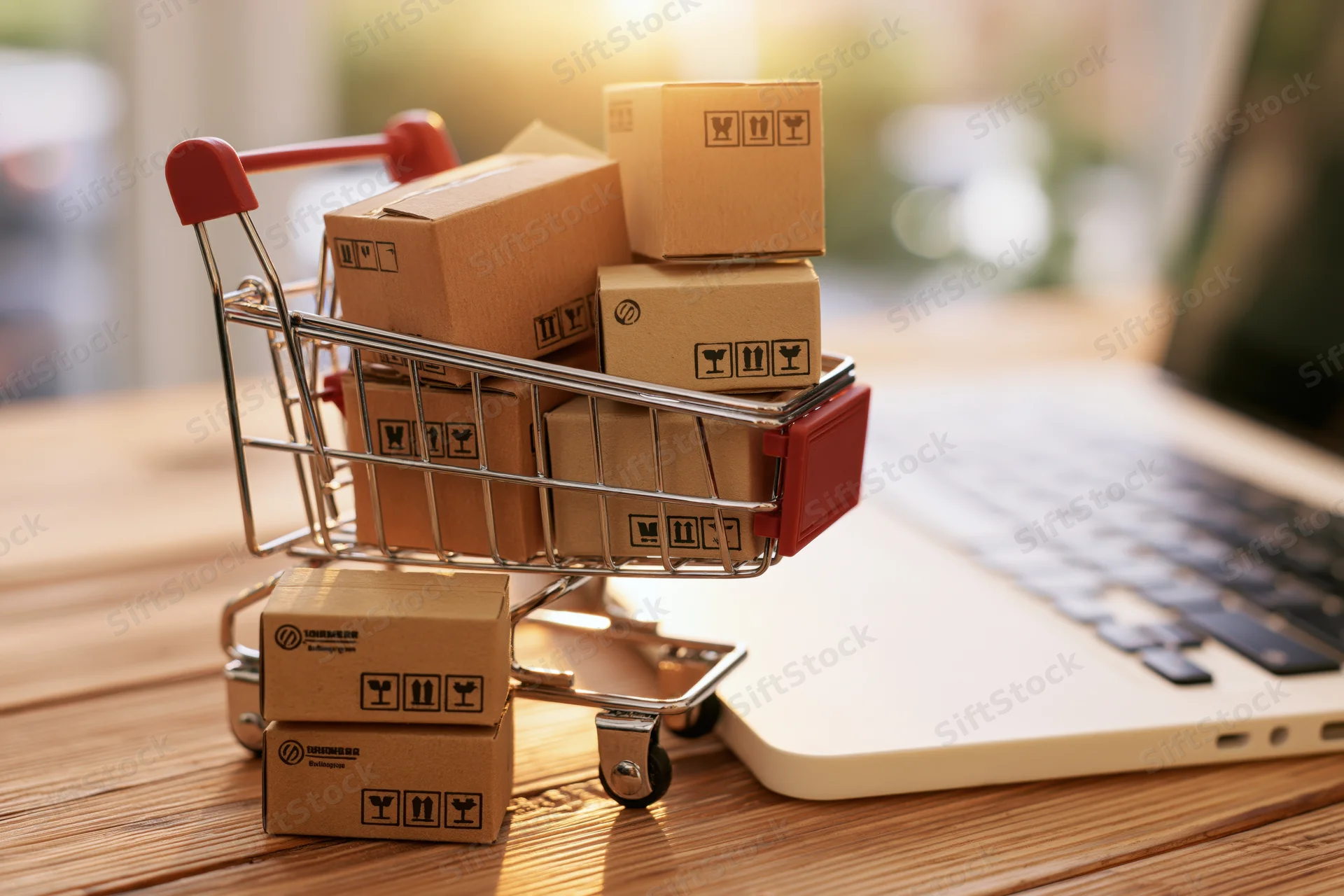 Miniature shopping cart filled with cardboard boxes on a desk beside a laptop