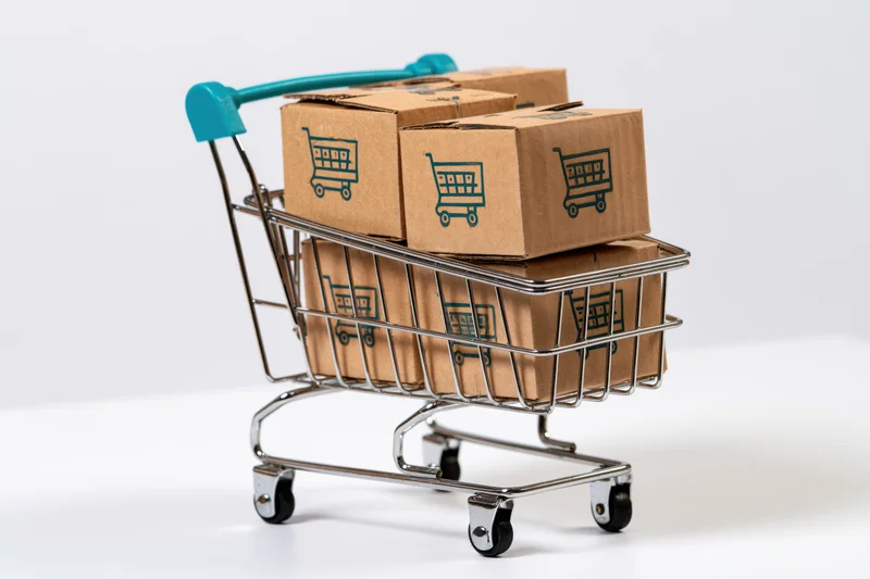 Miniature metal shopping cart filled with cardboard boxes printed with shopping cart icons on white background