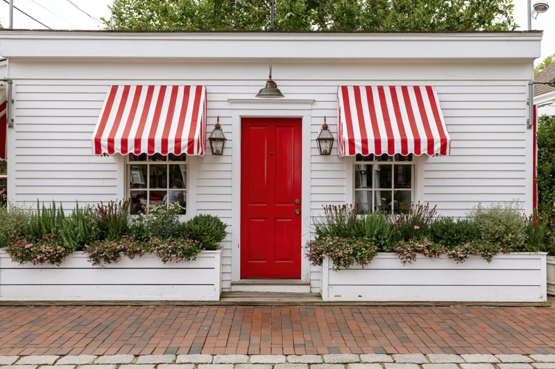 White cafe storefront with red door and striped awnings flanked by flower planters on brick sidewalk