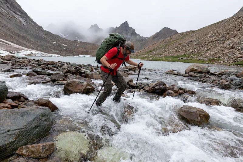 Male hiker crossing rocky mountain stream with trekking poles and backpack