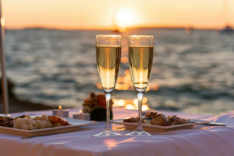 Two champagne flutes filled with sparkling wine on a white tablecloth by the ocean at sunset