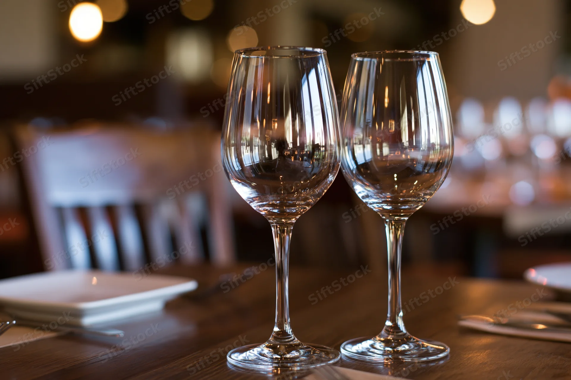 Two empty wine glasses on a dark wooden restaurant table with ambient lighting and table setting in background