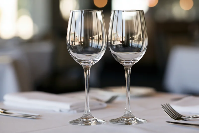 Two empty wine glasses on a white-linen restaurant table with silverware and napkins arranged for dining
