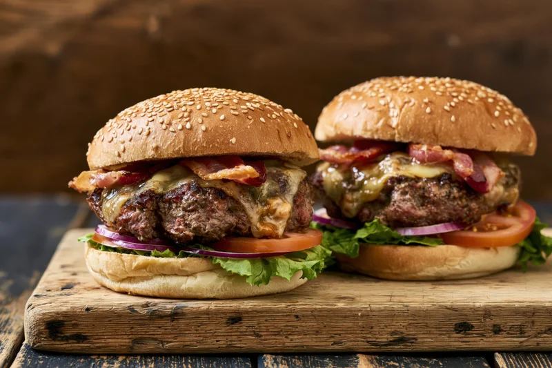 Two loaded beef burgers with cheese bacon lettuce tomato and onion on a worn wooden cutting board