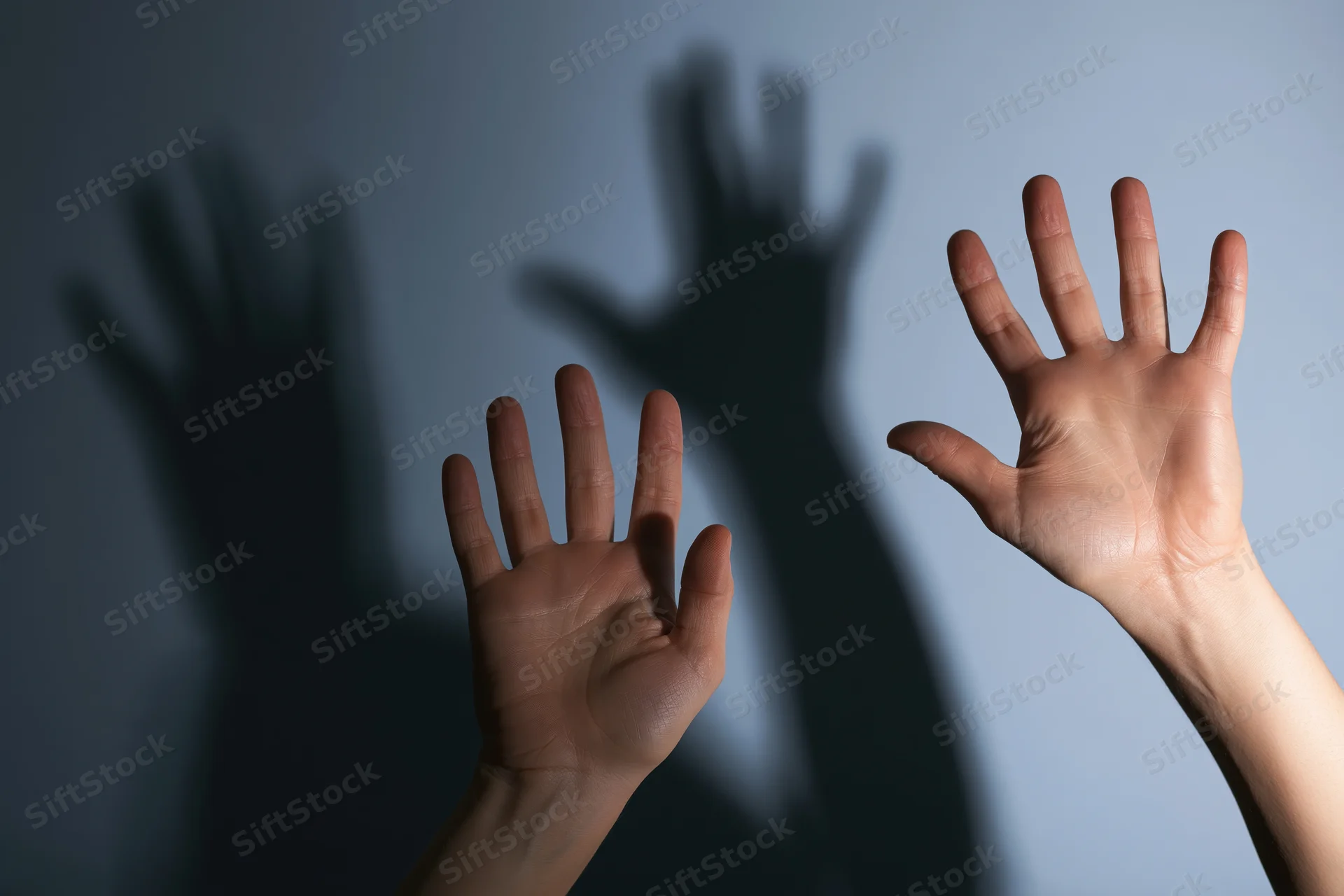 Two pale hands raised in a defensive blocking gesture against a shadowed wall