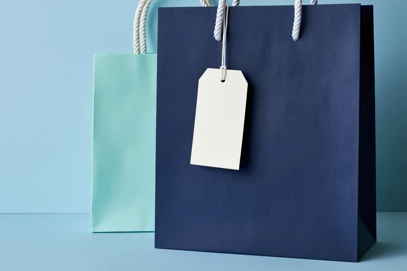 Two paper shopping bags side by side with a blank tag on the navy one against a blue background