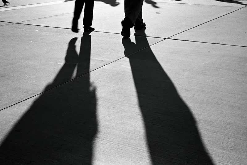 Two people walking away on a city sidewalk, their long shadows cast on the pavement