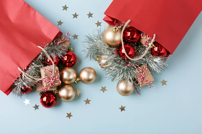 Two red paper bags spilling christmas ornaments and tinsel on a light blue surface