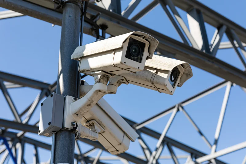 Two white cctv security cameras mounted on a metal pole against a blue sky with industrial framework in the background