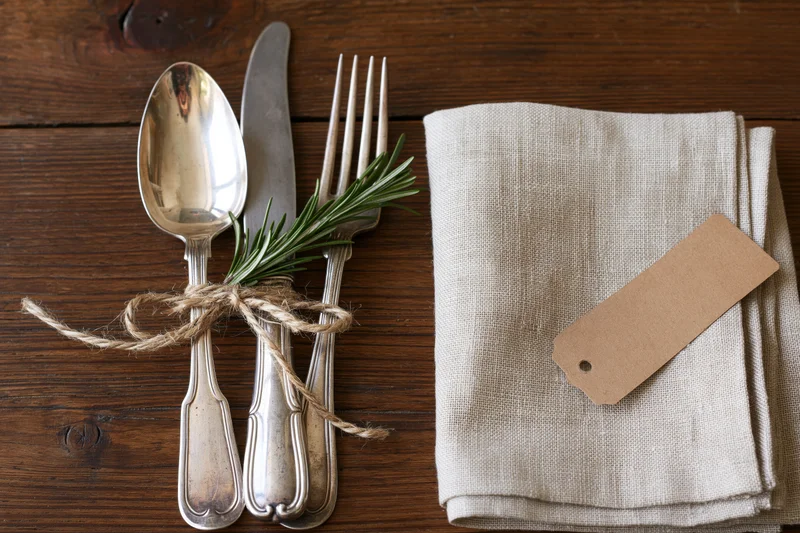Vintage silver cutlery bundle with rosemary and twine on wooden table beside linen napkin with paper tag