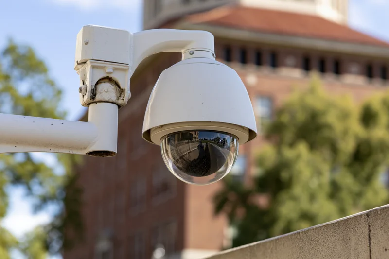 White dome security camera mounted on metal arm outdoors near brick building