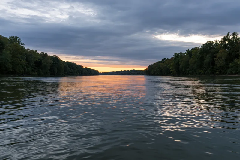 Wide river at dusk reflecting golden sunset light between tree-lined banks